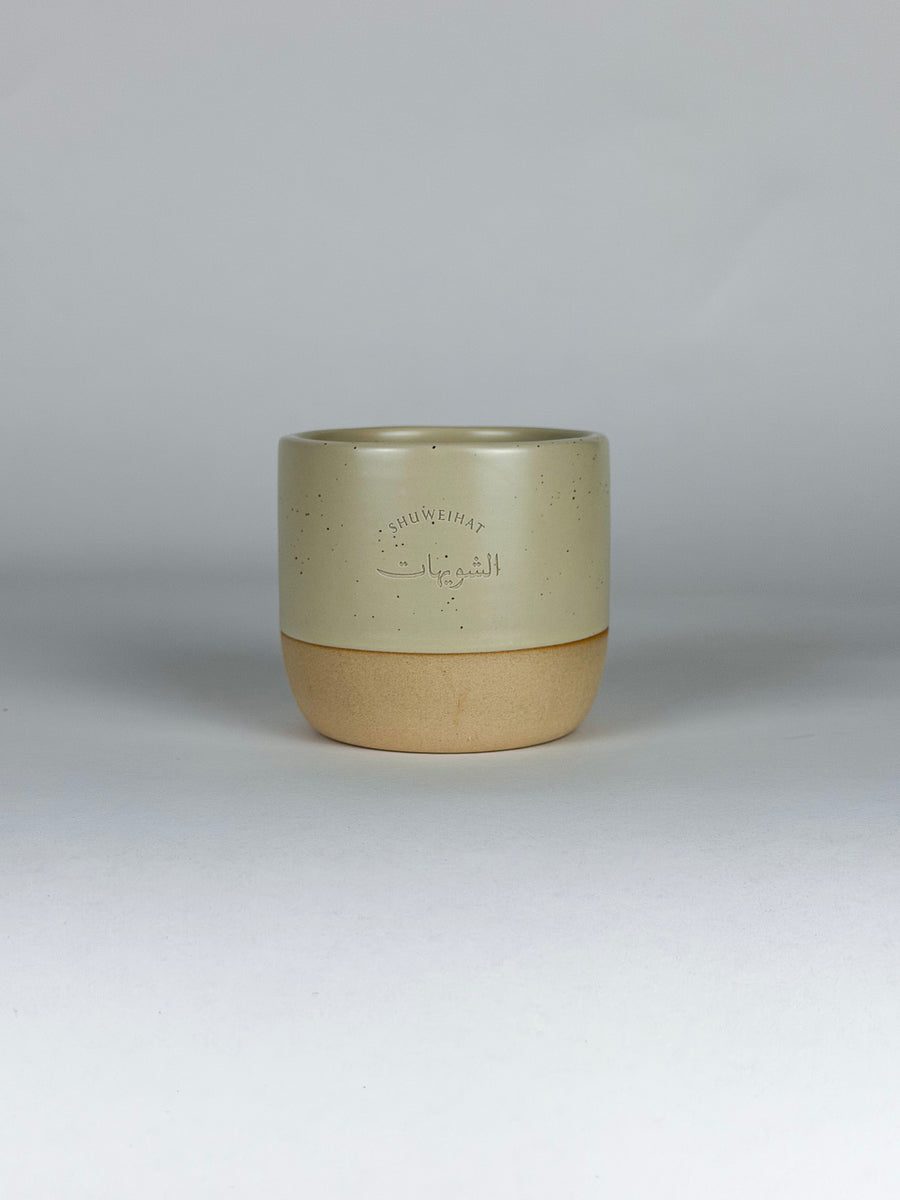 A ceramic cup with a two-tone design, featuring a speckled light beige upper half with embossed Arabic calligraphy and a solid tan lower half, set against a neutral gray background.